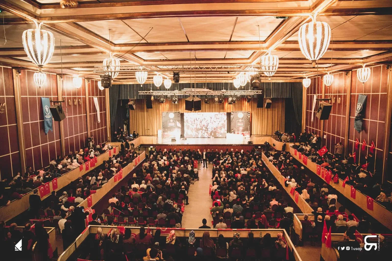 Festsaal mit Kronleuchtern, voll besetzten Tischen und Bühne